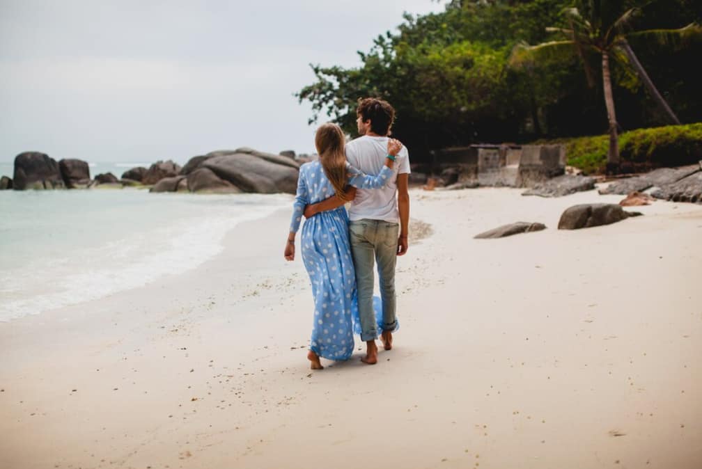 Two people stroll on a secluded beach with large rocks in the distance