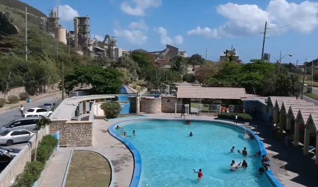 Swimmers and sunbathers at a crowded public pool, with industrial buildings in the distance